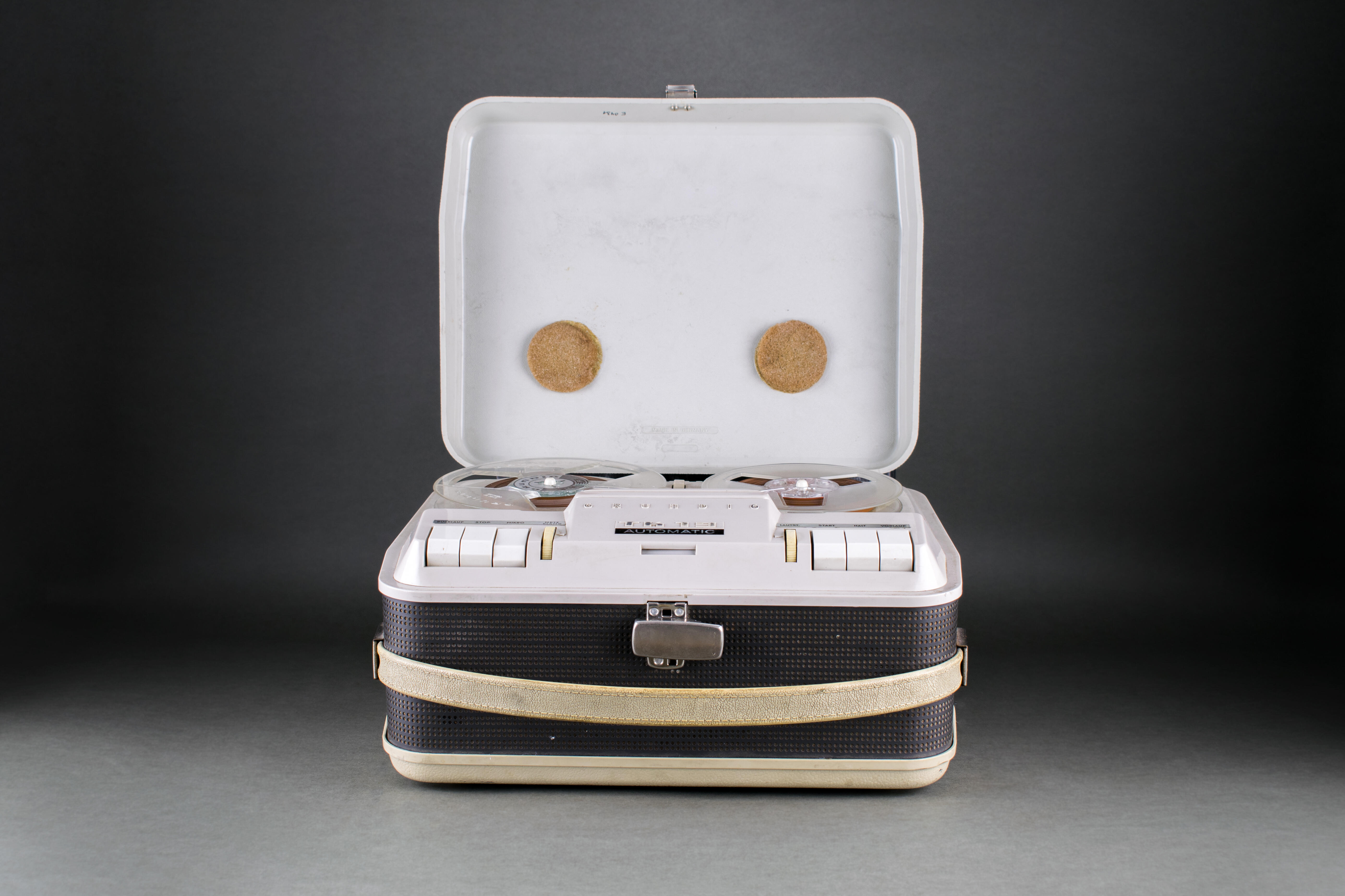 The picture shows a heavy small tape recorder from the 1960s with a removable grey lid, carrying handle and various buttons and controls.