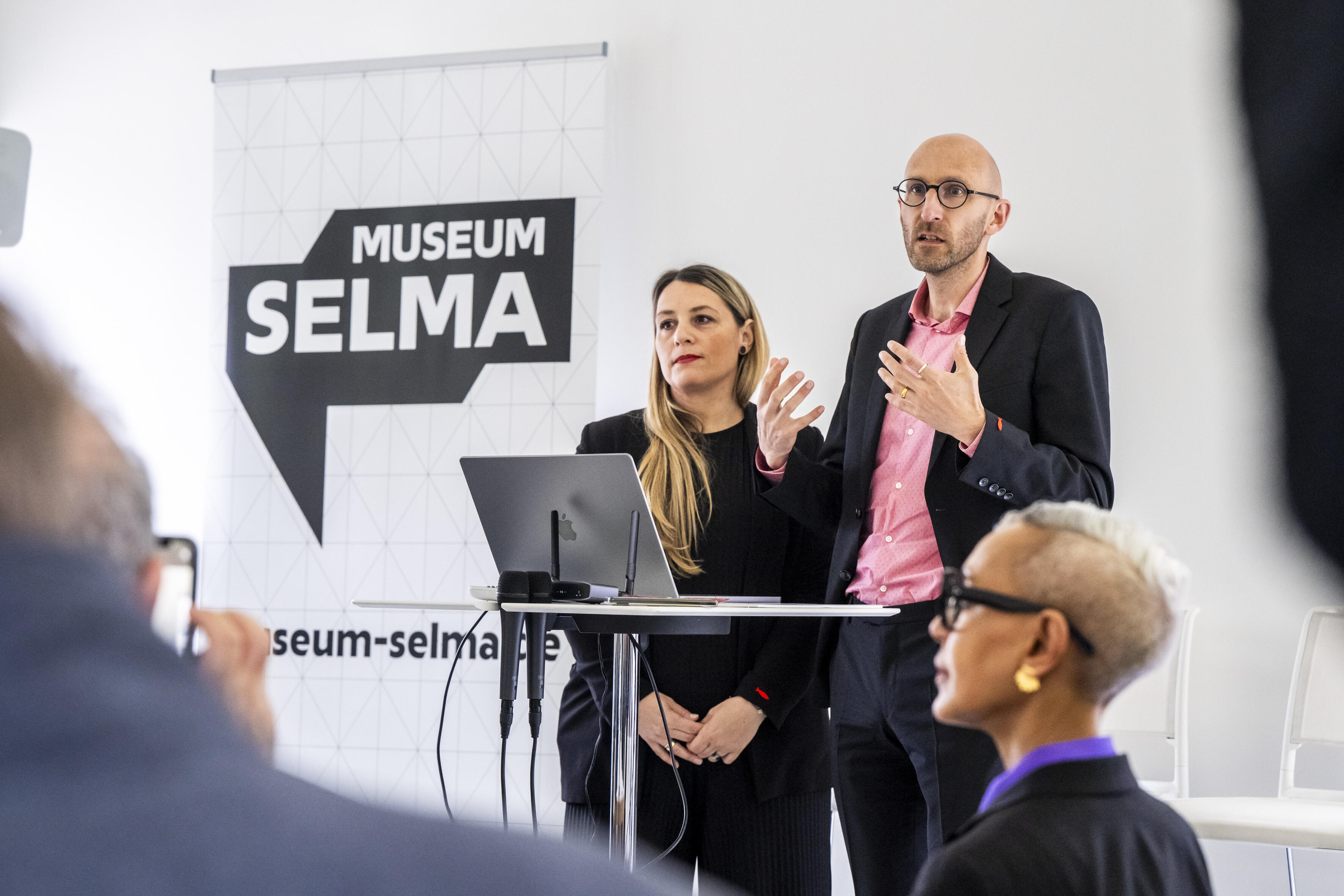 Zu sehen sind die Architekt*innen Michel Casertano und Alexandra Vassilakou von Atelier Brückner bei der Präsentation ihres Siegerentwurfes. Hinter ihnen steht das Logo von Museum Selma.