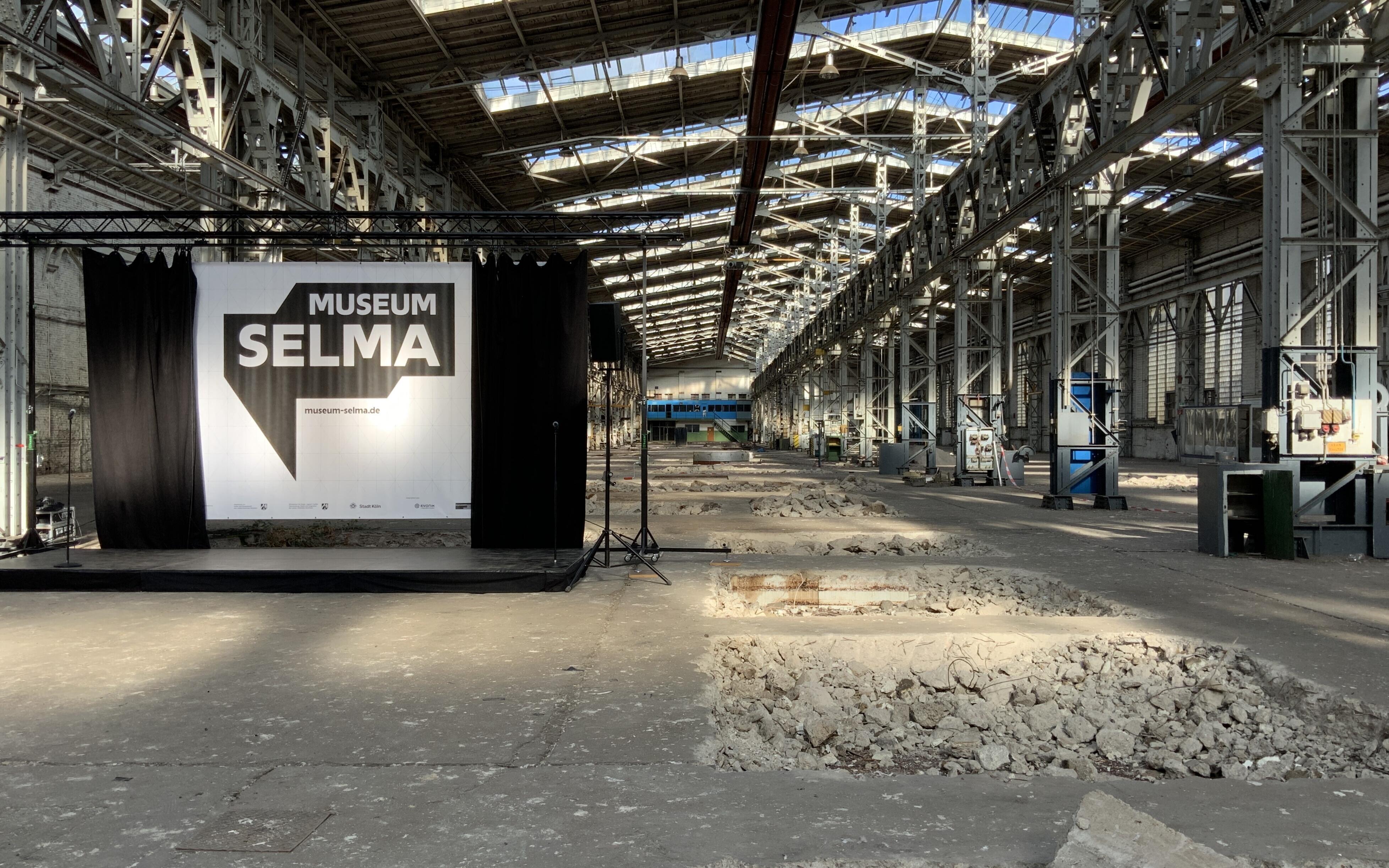Auf dem Bild ist eine alte Industriehalle zu sehen mit aufgerissenem Betonboden und hellem Lichteinfall aus den Oberlichtern des Hallendachs. Am linken Bildrand ist eine Bühne aufgebaut und ein schwarzer Vorhang geöffnet. Dahinter ist das Logo von Museum Selma zu sehen.