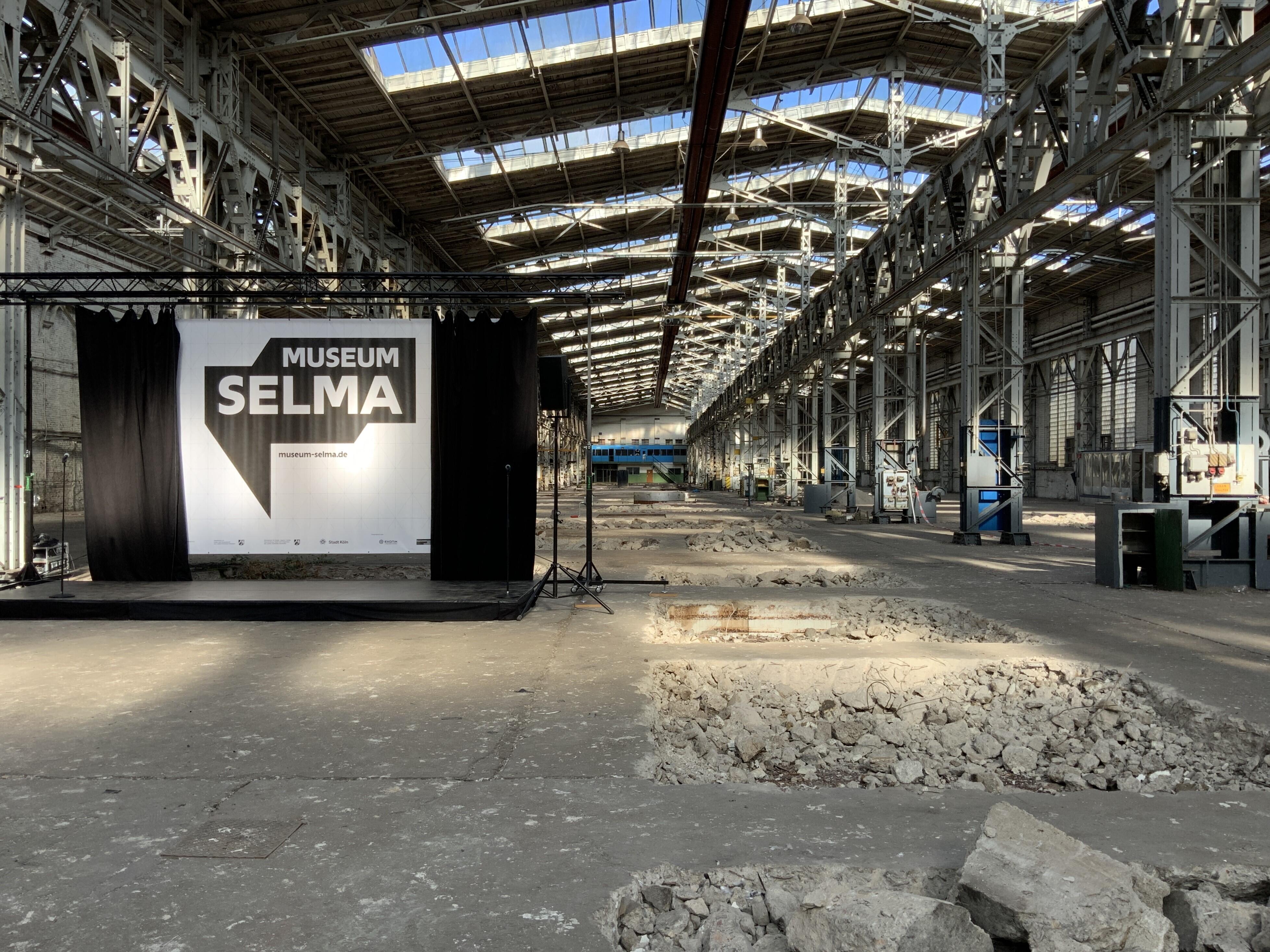 Auf dem Bild ist eine alte Industriehalle zu sehen mit aufgerissenem Betonboden und hellem Lichteinfall aus den Oberlichtern des Hallendachs. Am linken Bildrand ist eine Bühne aufgebaut und ein schwarzer Vorhang geöffnet. Dahinter ist das Logo von Museum Selma zu sehen.