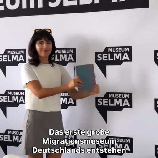 Ein Foto von Autorin Seyda Kurt vor einer Museum Selma Pressewand. Sie präsentiert ihr Notizbuch.